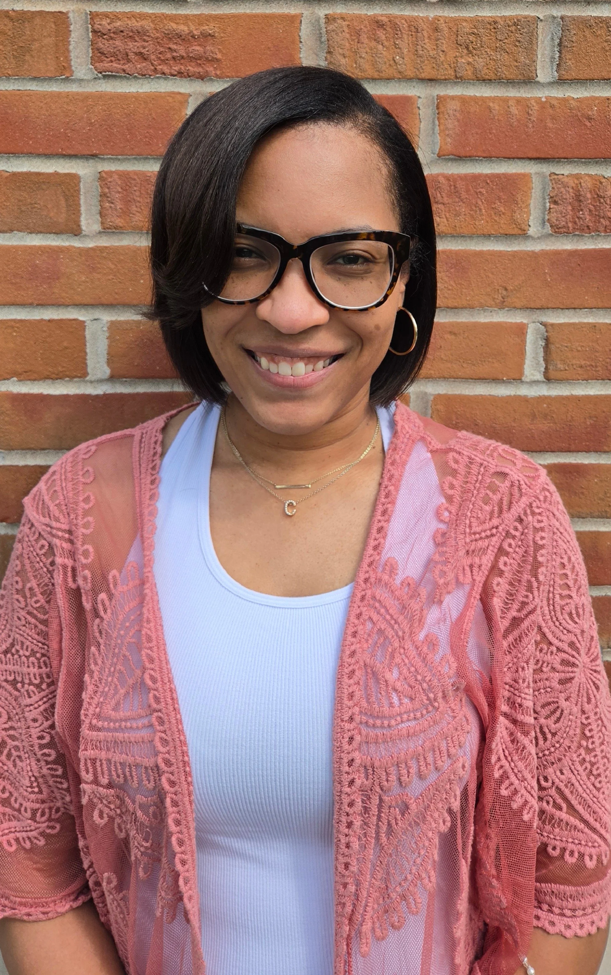 Smiling woman with glasses and short curly hair standing in front of a brick wall.
