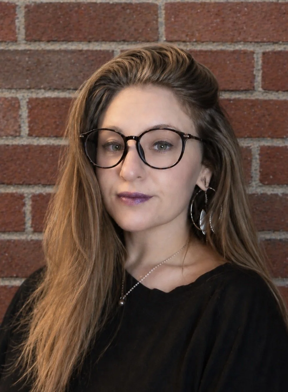 A woman with wavy blonde hair, glasses, and multiple face piercings smiling against a brick wall.