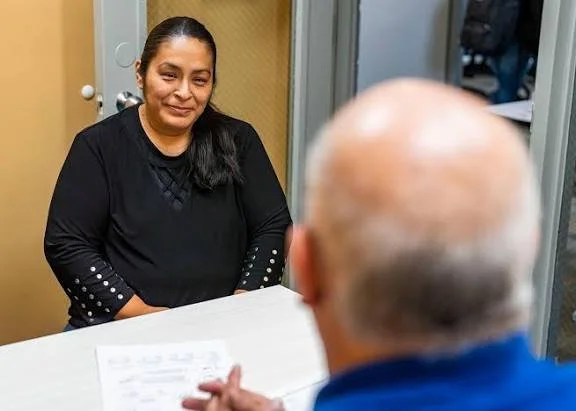 A woman with long dark hair sitting at a table across from a person with short white hair, seen from behind. The setting appears to be an office or consultation room with a door and windows in the background.