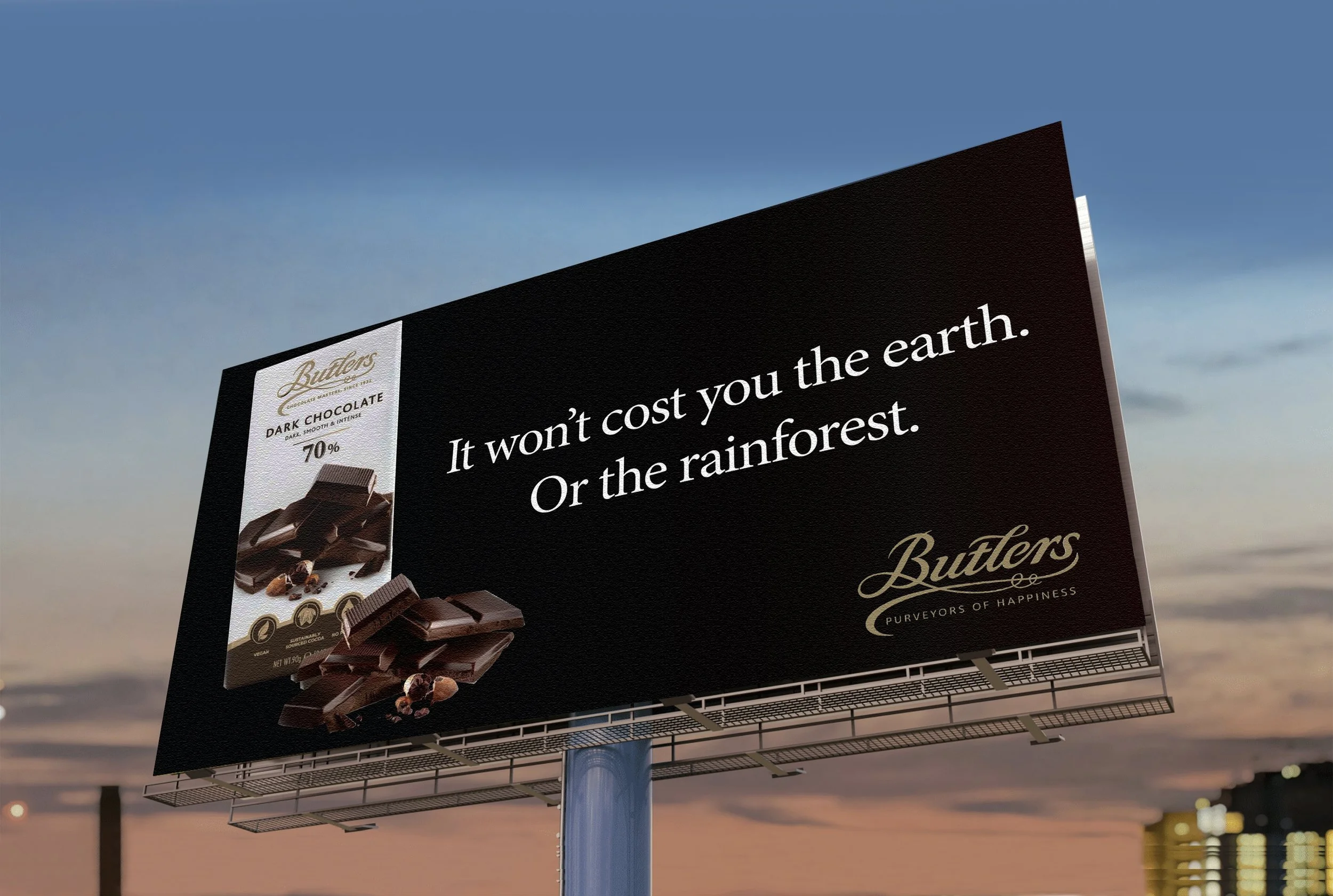 Billboard advertisement for Butlers dark chocolate. The billboard features an image of broken dark chocolate bars with almonds. The text on the billboard says, "It won't cost you the earth. Or the rainforest." The background is a sunset sky with city