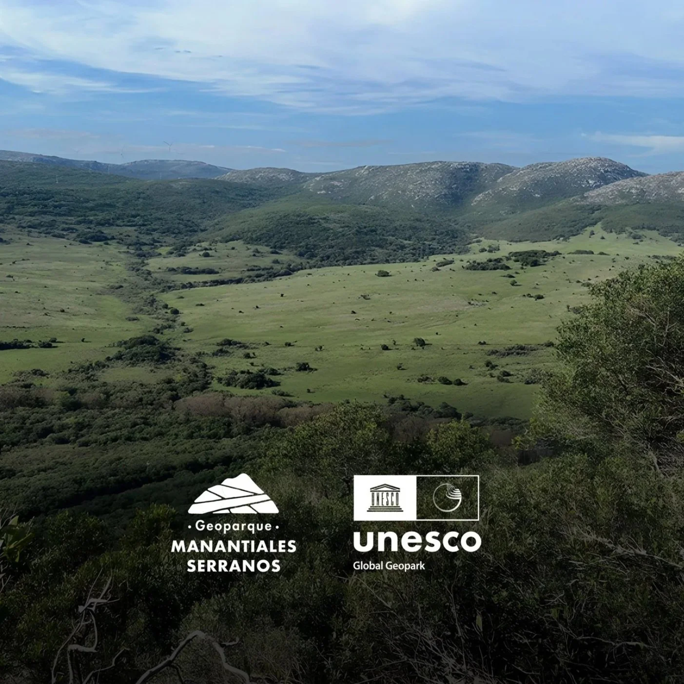 Paisaje de colinas verdes y montañas en un parque geológico llamado Manantiales Serranos, con un cielo parcialmente nublado.