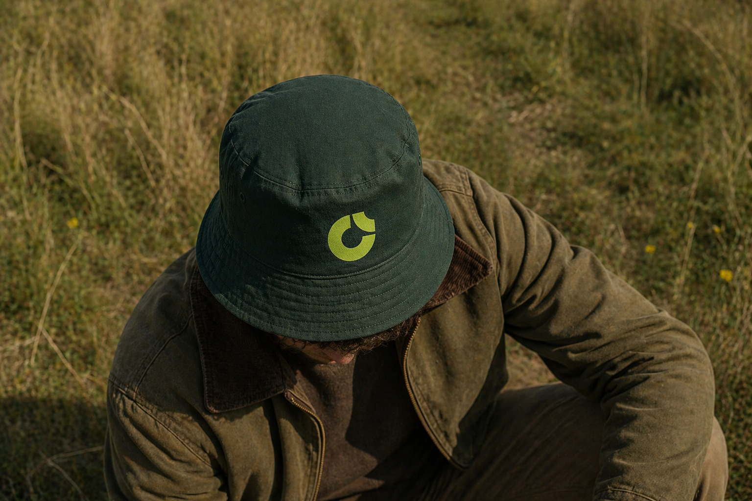 Persona con sombrero verde con logotipo amarillo, vestido con chaqueta marrón y pantalones en campo de hierba.