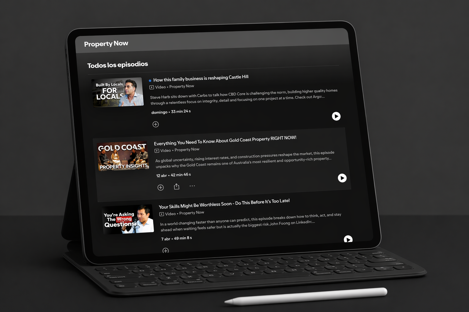 Pantalla de una tableta con una lista de episodios de un servicio de streaming, mostrando miniaturas, títulos, descripciones y controles de reproducción, con un teclado y un stylus en un fondo oscuro.