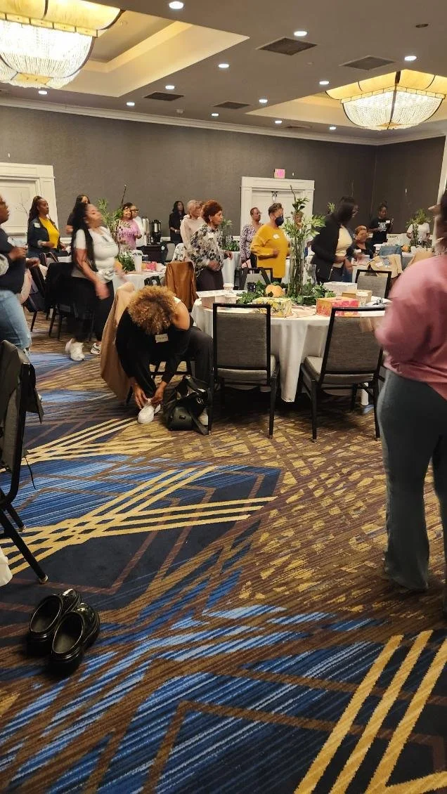 A group of women gathered in a banquet hall, some standing and some seated around decorated tables, with a woman bending down to tie her shoe.