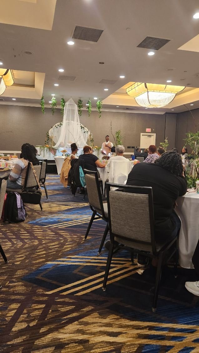 People seated at round tables in a banquet hall, watching a person speak at a decorated stage with greenery and a canopy, in a formal event setting.