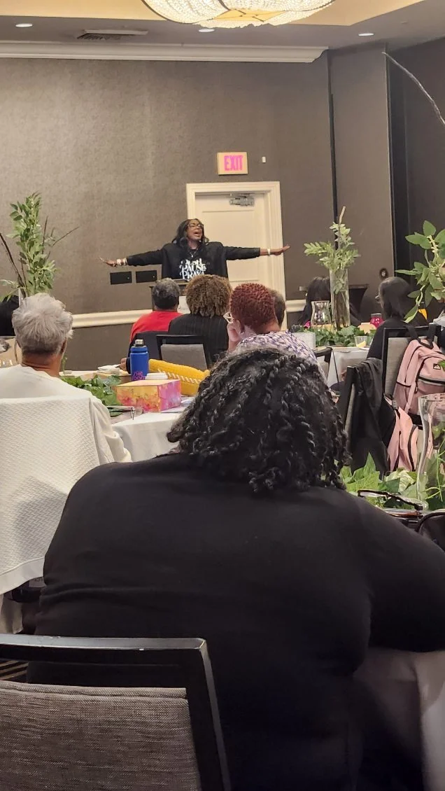 A woman standing at a table with her arms outstretched, speaking to an audience at a banquet or conference in a decorated room.
