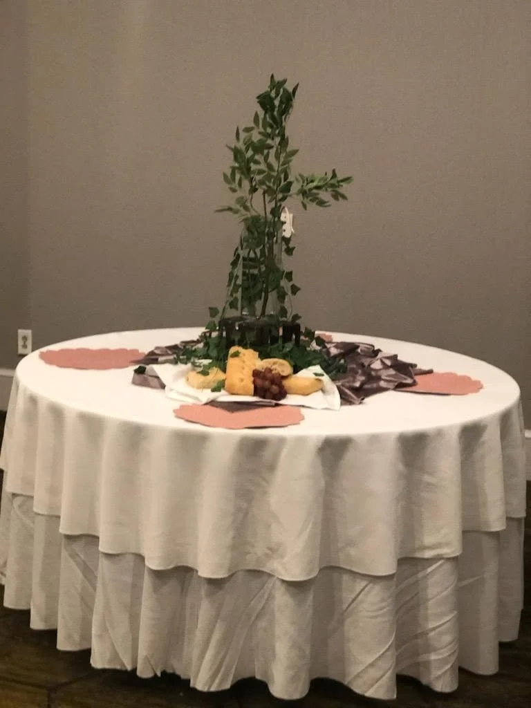 A round banquet table with a white tablecloth and decorative pink and purple paper placemats. The centerpiece features a small tree or shrub with leaves and some food, including cheese, grapes, and crackers, arranged around the base.