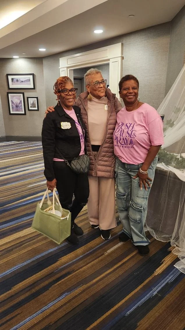 Three women standing together indoors, smiling, with a wall and framed pictures in the background. Two women on the left and right are wearing pink shirts, and the woman in the middle is wearing a puffer jacket and light-colored pants.