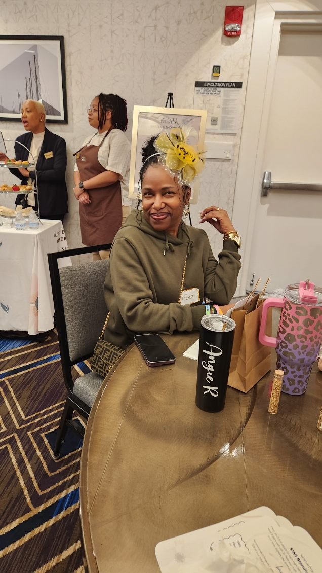 Woman sitting at a table with a festive yellow and black fascinator hat, smiling. Two women are standing in the background, one in a uniform, the other in a black blazer. There are gift bags, a tumbler with 'Angel R' written on it, and a sippy cup on
