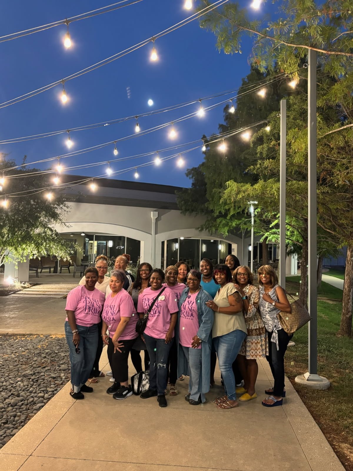 A group of women standing outdoors at night in front of a building, illuminated by string lights overhead, with trees and a clear evening sky visible.