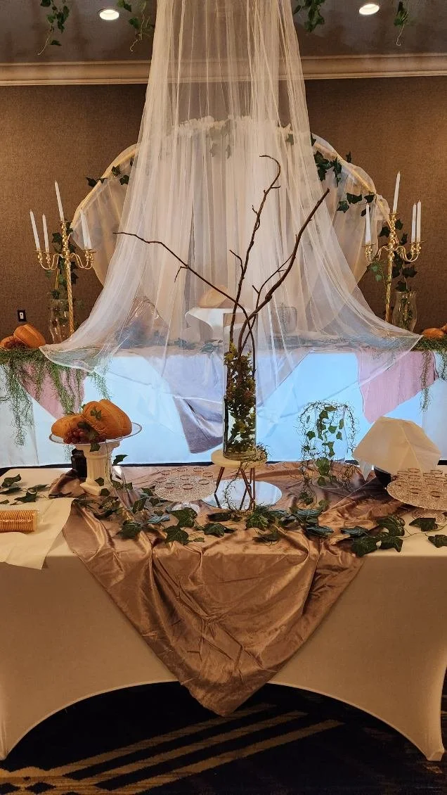 Table decorated with beige and pink tablecloth, featuring autumn pumpkins, greenery, and candles, with a sheer white canopy and branches as part of a centerpiece, likely for a wedding or special event.