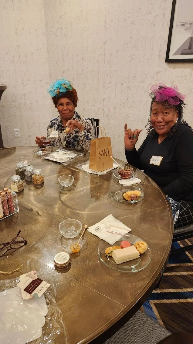 Two women wearing colorful fascinators sitting at a round table with desserts and small jars of sprinkles, laughing and making gestures, in a room with a patterned wall and framed artwork.