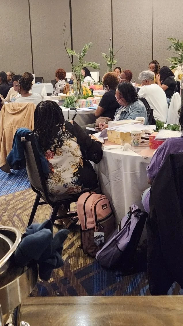 A group of women sitting at tables during a conference or workshop in a large room with beige walls, some with laptops, notebooks, and cups, engaged in listening or taking notes.