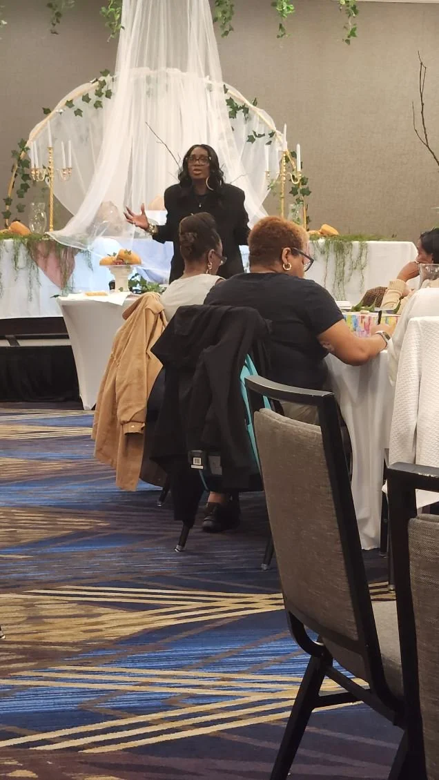 Woman giving a speech or presentation at a decorated event with tables and guests listening.