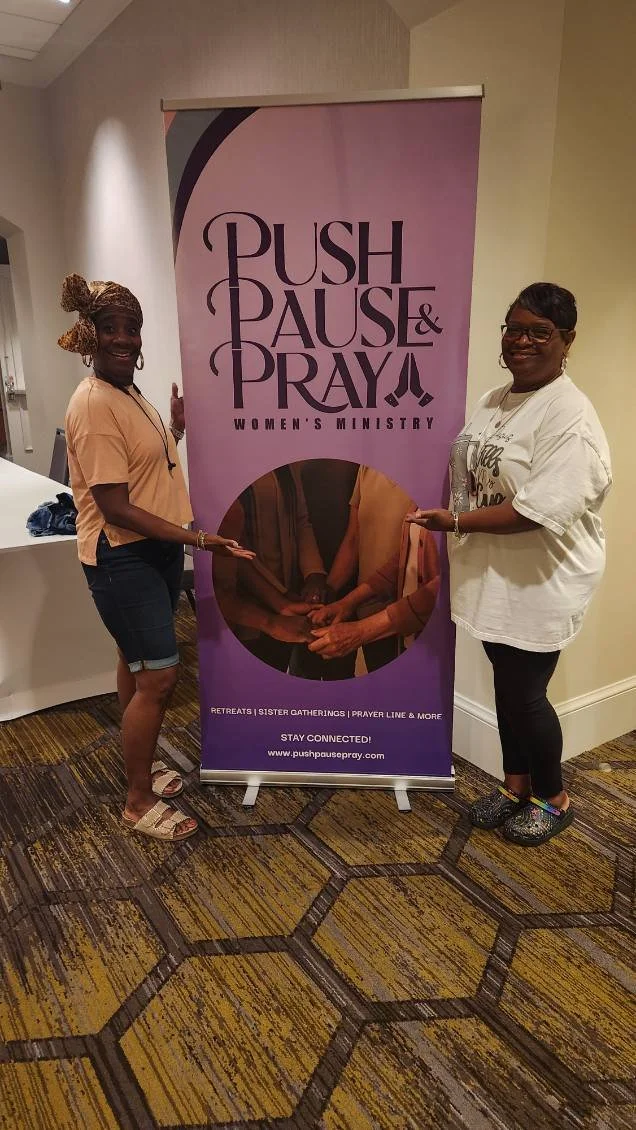 Two women standing beside a vertical banner that reads "Push Pause & Pray Women's Ministry." There is a circular photo on the banner showing hands holding each other, and additional text indicating retreats, sister gatherings, prayer line, and a webs