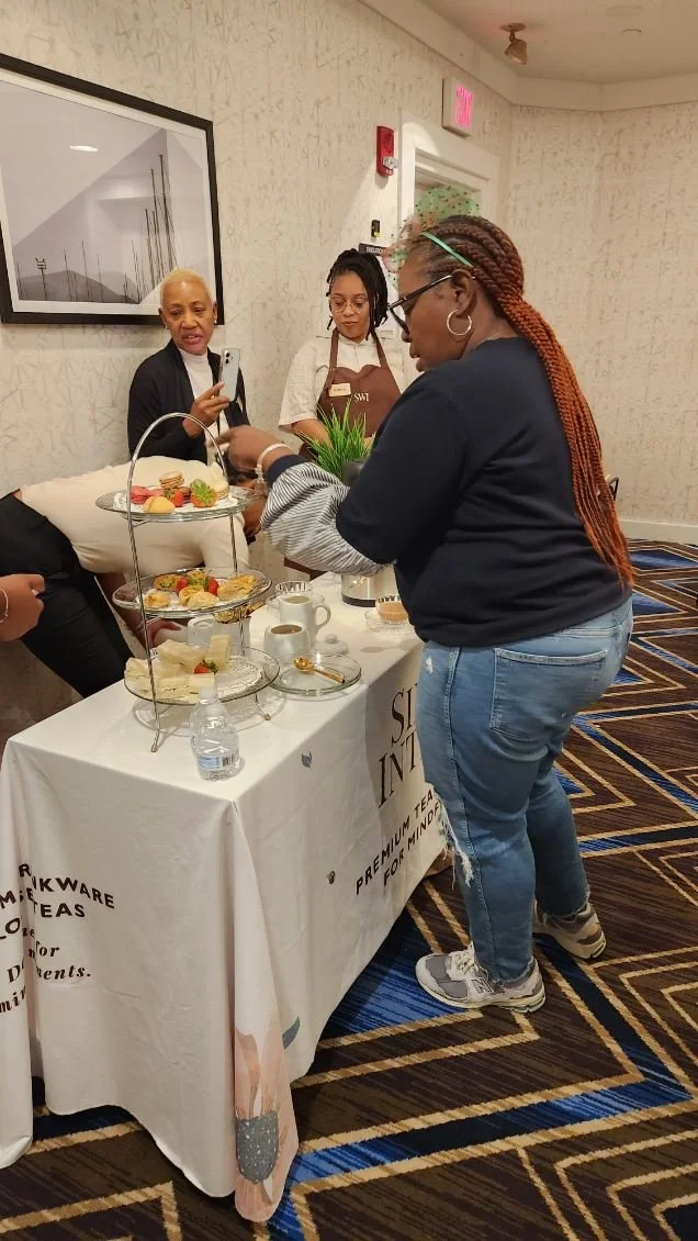 Three women are gathered around a dessert table at an indoor event. One woman with long braided hair and glasses is taking a photo or video, while another woman with short blonde hair looks on. The third woman, with glasses and a brown apron, stands 