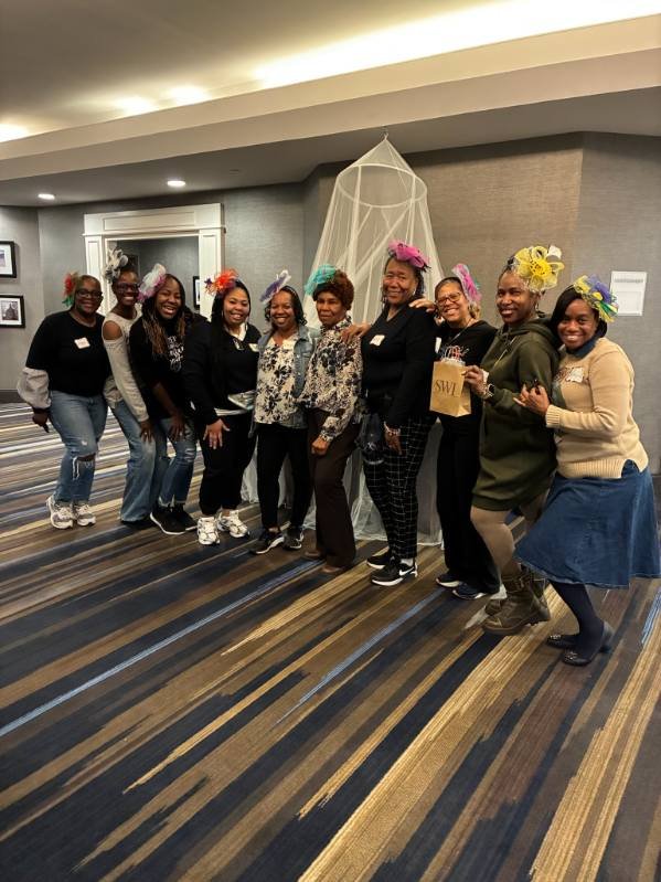 A group of nine women standing in a line indoors, all wearing colorful flower headbands. They are smiling and posing for the photo in front of a wall with a decorative net structure hanging from the ceiling.