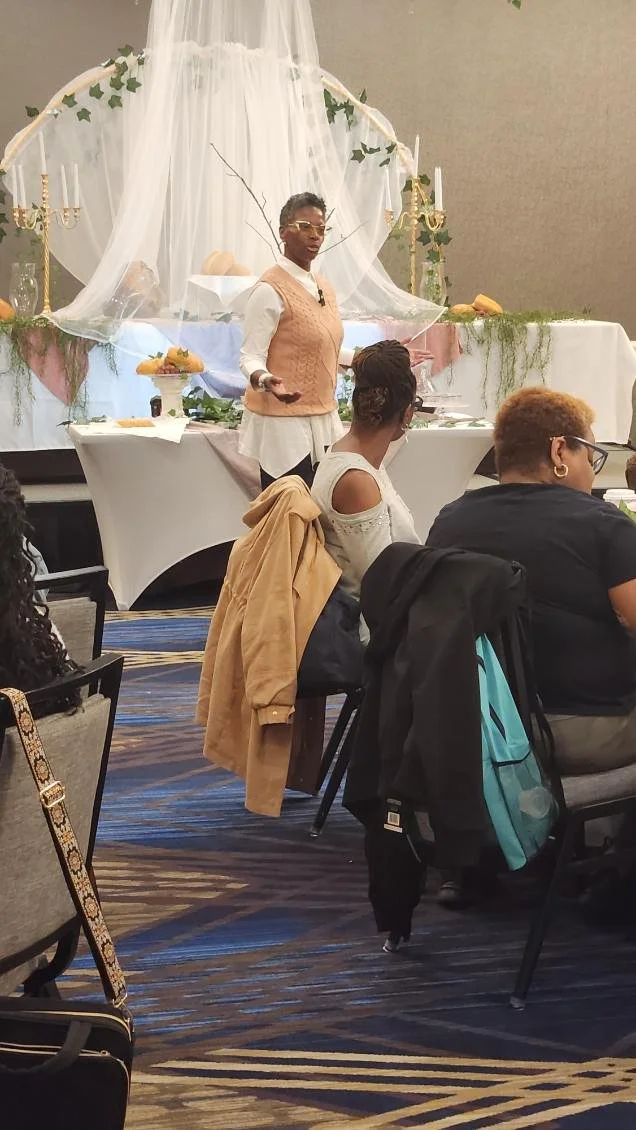 A woman is giving a presentation or speech at a decorated event table with white drapery, candles, greenery, and gourds, while several people sit attentively facing her.