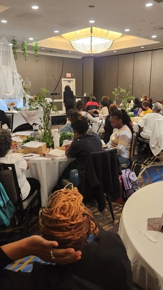 A woman with orange dreadlocks sitting at a table in a conference room, holding her hair. There are many attendees seated at round tables, facing a stage with a presenter. The room has a large chandelier ceiling light and is decorated with greenery.