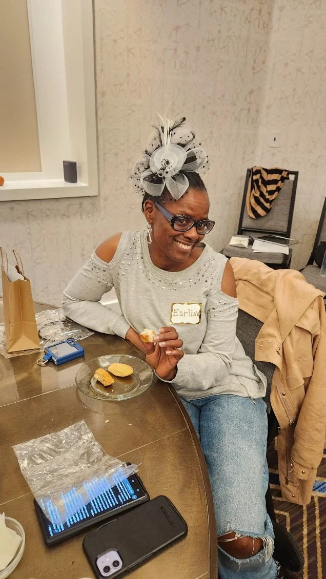 A woman sitting at a table with a smile, wearing glasses, a gray cold-shoulder sweater with pearl embellishments, and a decorative black and white fascinator in her hair. She has a name tag that says "Earlia" and is holding a biscuit or cookie. The t