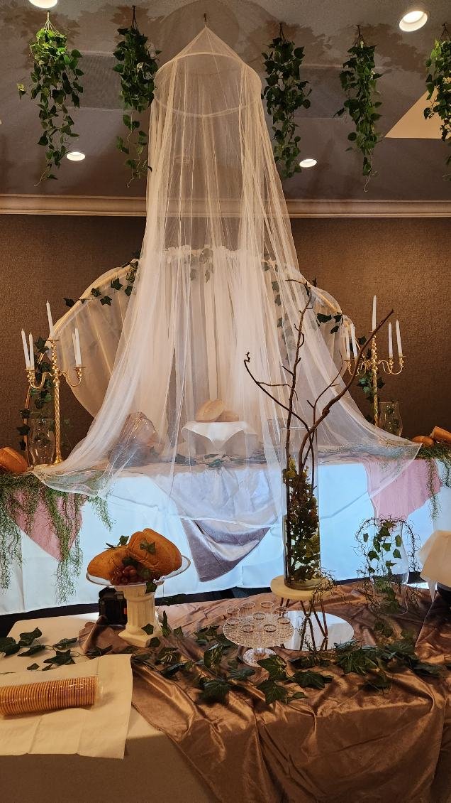Table decorated with greenery, candles, and a branch in a glass vase, with a white cake and carved pumpkins in the background, set for a fall celebration.