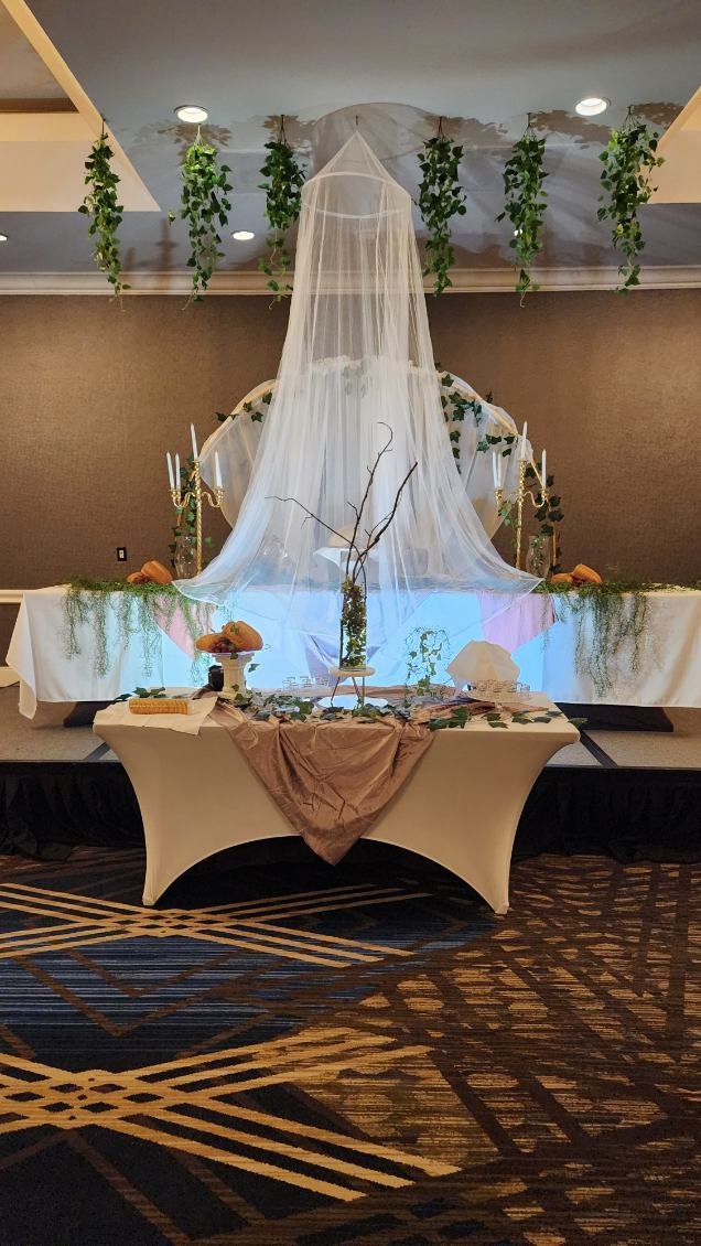 Decorative table setup with draped cloth, a branch centerpiece, white candles, and green plants hanging overhead, suggesting a wedding or special event.