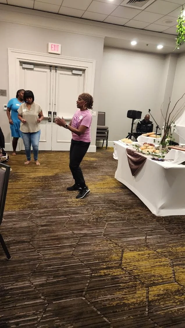 A woman in a pink T-shirt and black pants dancing in a banquet hall with other women and a man in the background near a buffet table.