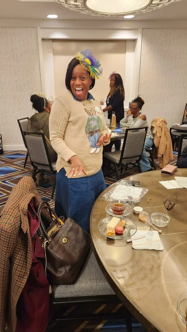 A woman wearing a beige sweater with a graphic of a child and a colorful hat stands at a buffet table, smiling and gesturing. Around her are several women seated at chairs, with an elegant ceiling, patterned carpet, and a round table with food and ut