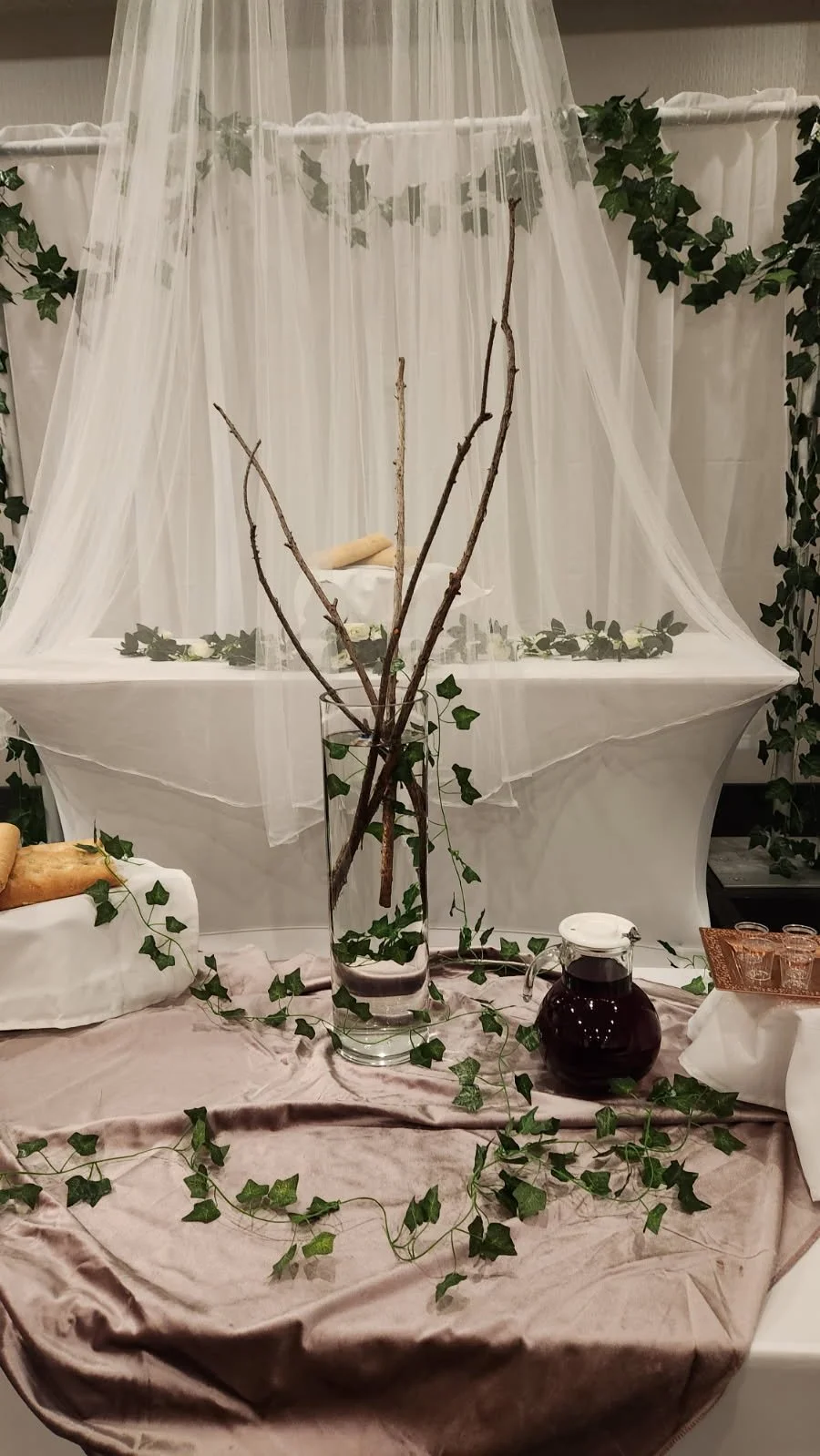 Display of a table with a tall glass vase holding bare tree branches, decorated with faux ivy vines. In the background, there is a white canopy with sheer fabric and green ivy vines, creating a decorative backdrop. The table is covered with a light p