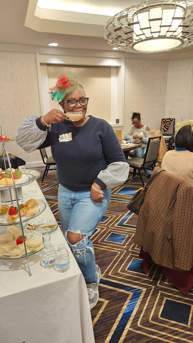 A woman smiling and posing with a cup of tea or coffee at a social gathering or buffet table in a banquet hall. She is wearing glasses, a navy sweater with striped sleeves, and ripped jeans. The hall has a patterned carpet, a large ceiling light, and