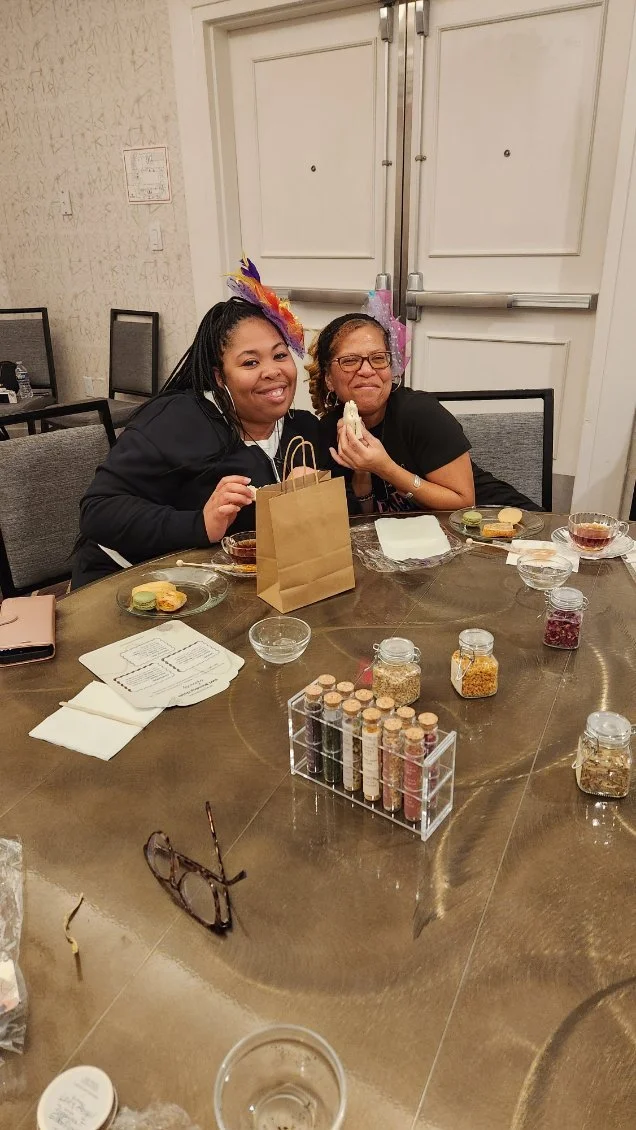 Two women sitting at a round table, smiling and holding food, with various jars and plates of snacks and seasonings on the table, and a closed door in the background.