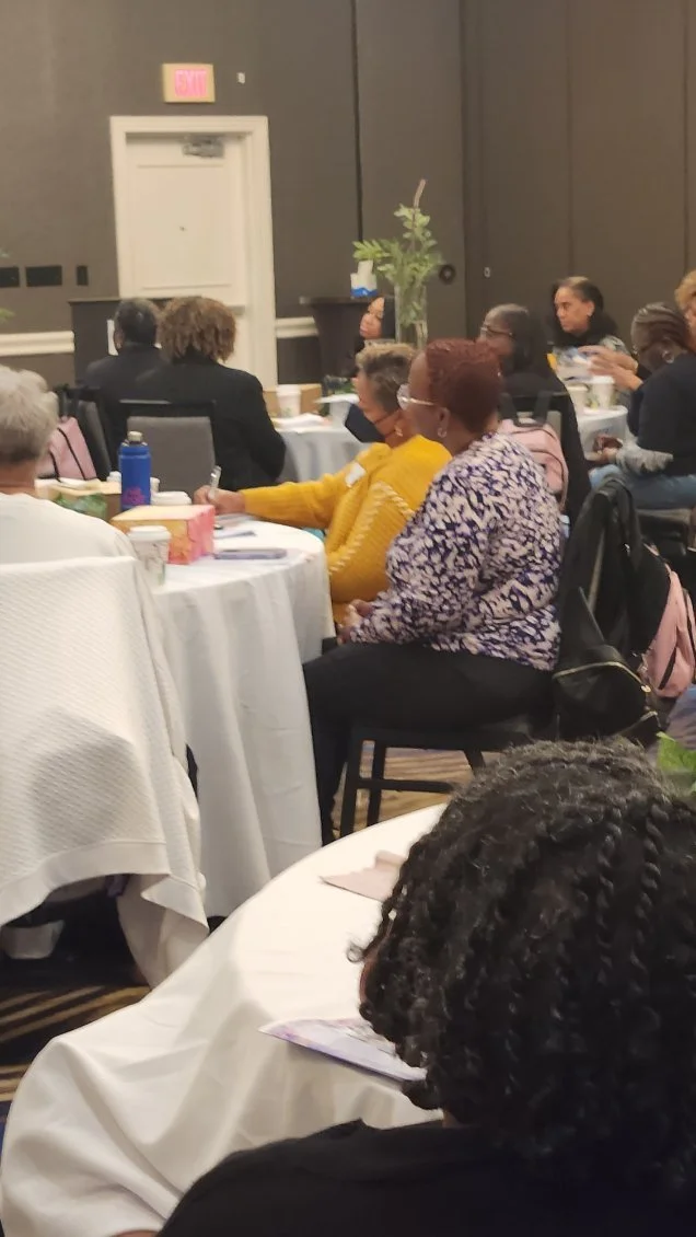 Women attending a conference or meeting seated at round tables with white tablecloths, some with coffee cups and notepads, in a dimly lit room with dark walls, plants, and a door in the background.