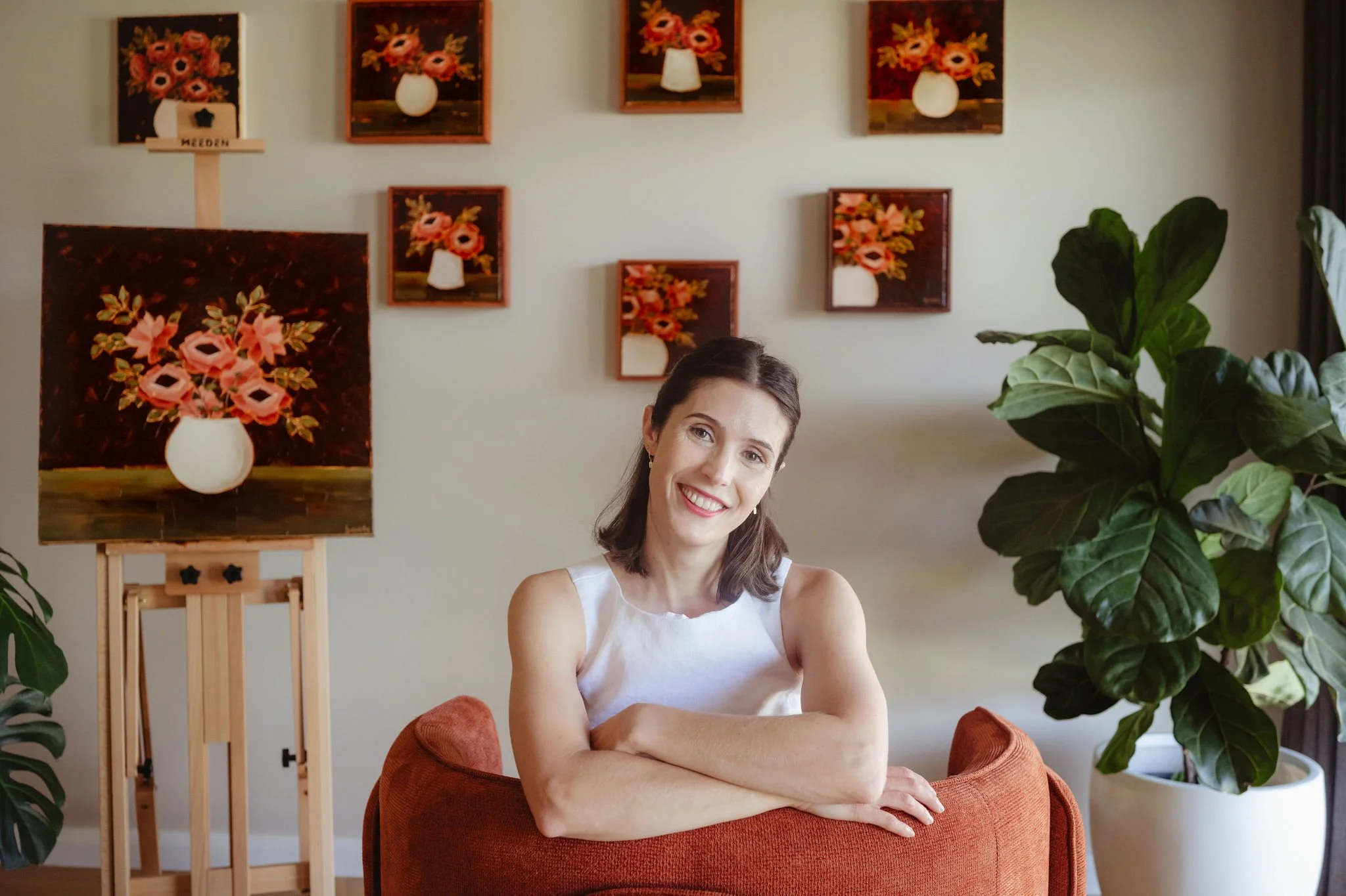 Chloe Boots Australian oil painter sits on a red armchair with her original floral paintings on the wall and green plants nearby.