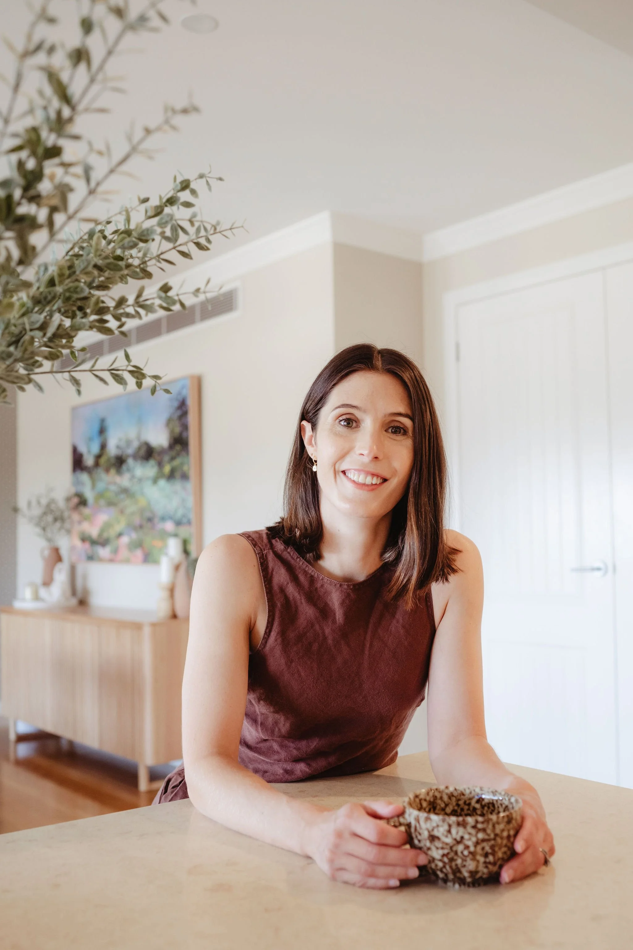 A woman with shoulder-length brown hair, wearing a sleeveless brown shirt, smiling while sitting at a kitchen counter with a ceramic mug in her hands. The background features a light-colored wall, a painting, and a wooden sideboard with decorative items.