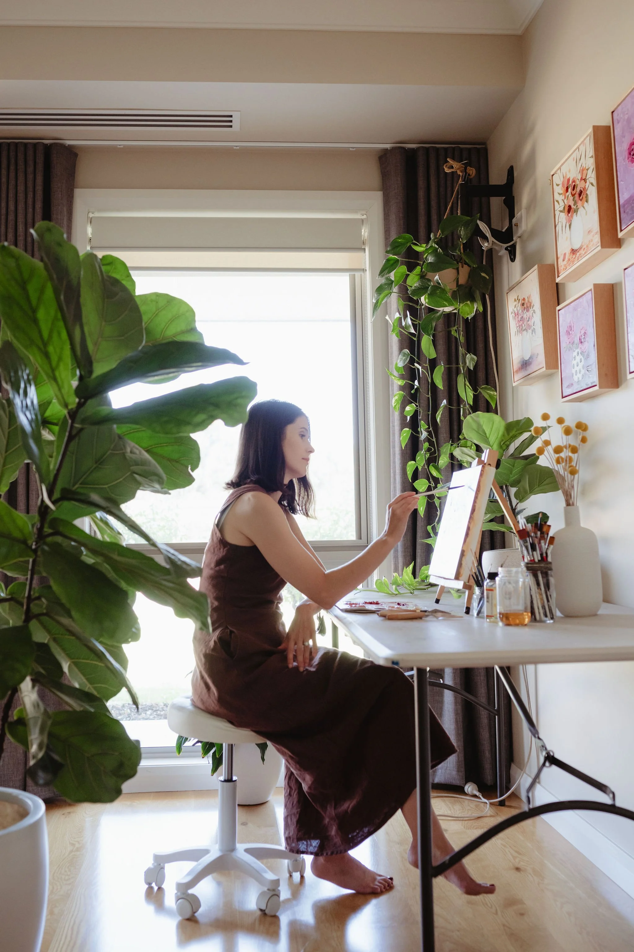 Woman sitting at a white desk, painting or drawing on a canvas, surrounded by houseplants and wall art, with a window in the background.