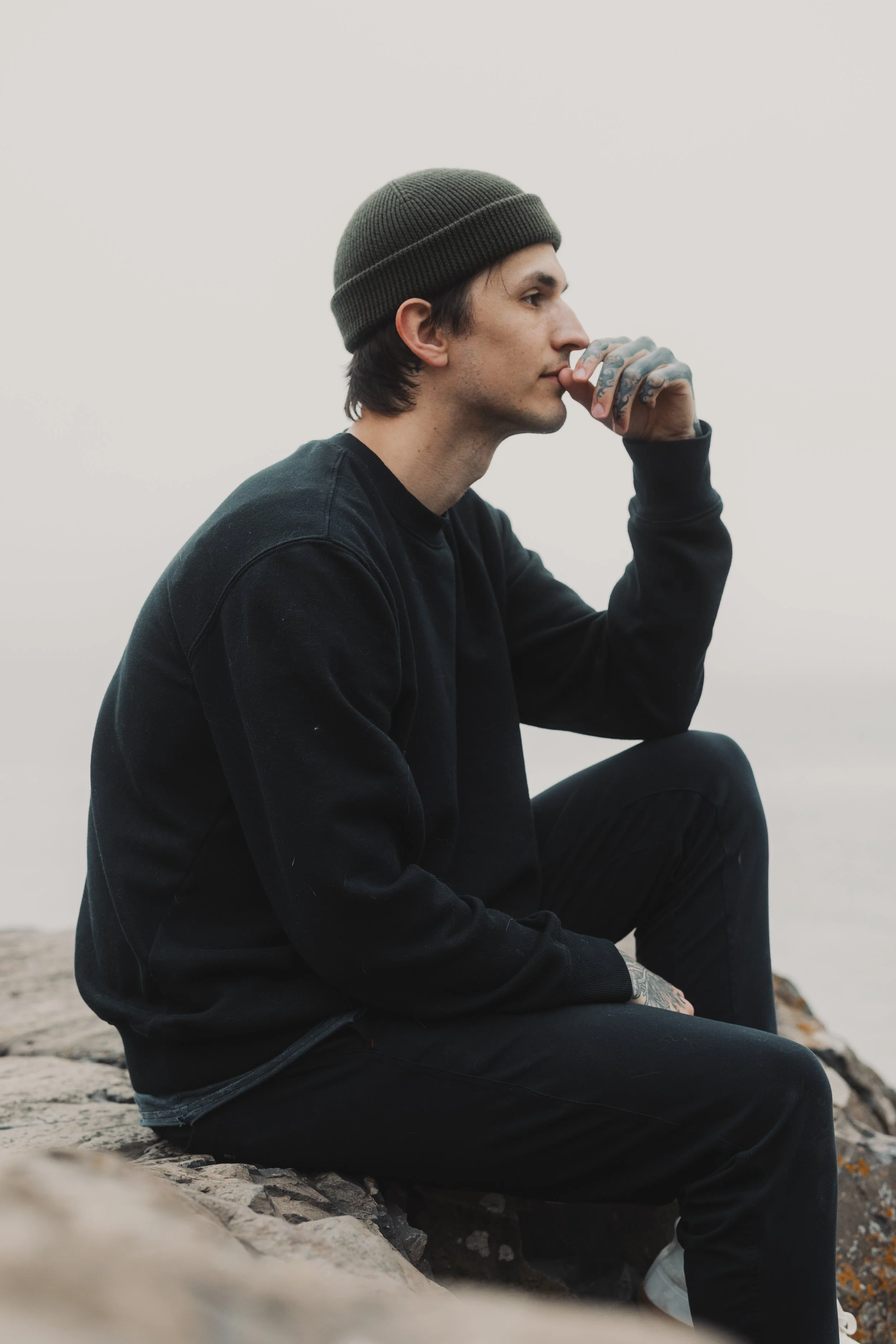 A young man sitting on rocks near the water, wearing a green beanie, black sweatshirt, and black pants, with tattoos on his hands, looking to the side with a thoughtful expression.