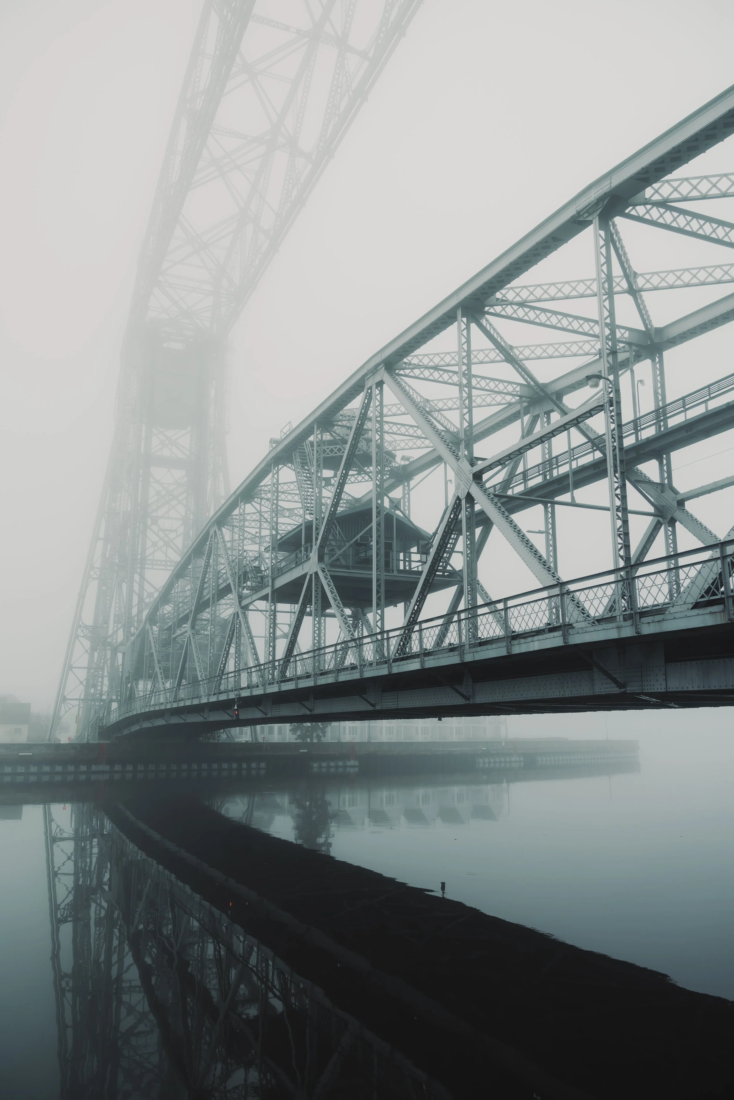 A foggy view of a large steel bridge over water with reflections, featuring detailed structural beams and a small control room.