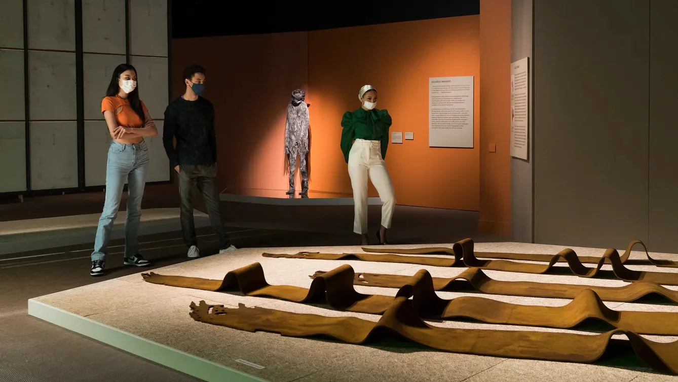 Photo of the exhibition installation for Rooted Beings at Wellcome Collection. There are 3 people standing around a large kelp sculpture in a dim room with spot lighting.