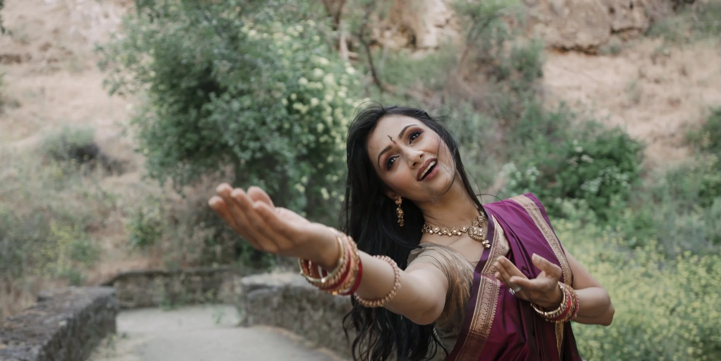 A woman dancing outdoors in traditional Indian attire, wearing a purple saree, bangles, and jewelry, with trees and foliage in the background.
