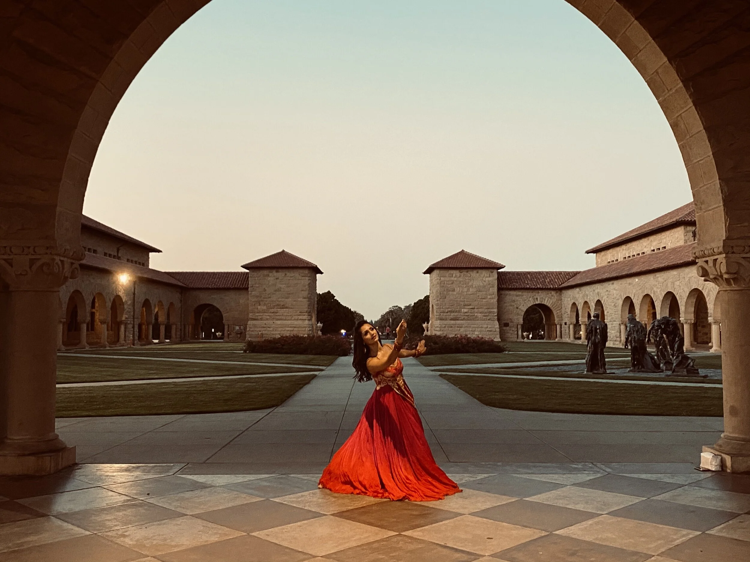 A woman in a vibrant red dress dancing under an archway in a courtyard with stone buildings and sculptures in the background.