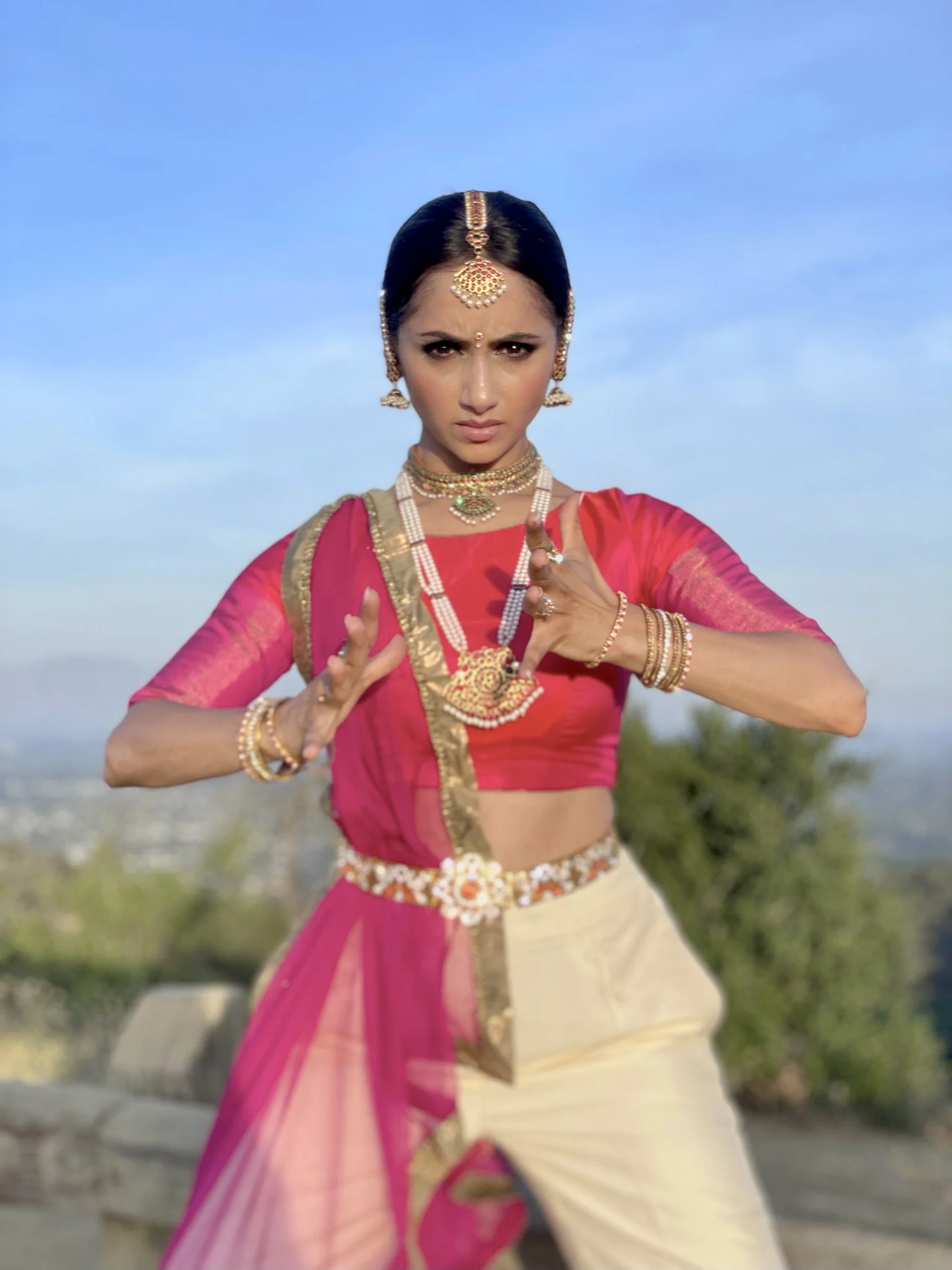 Woman in traditional dance attire performing a pose outdoors, wearing pink and gold garments with jewelry.
