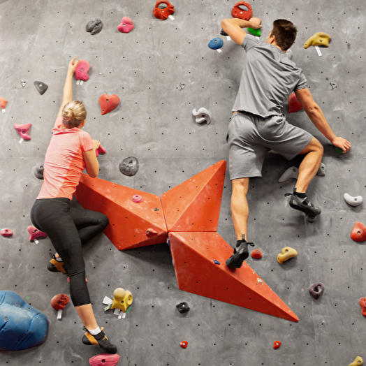 Two people rock climbing on an indoor gym wall with colorful holds and a large orange volume feature.