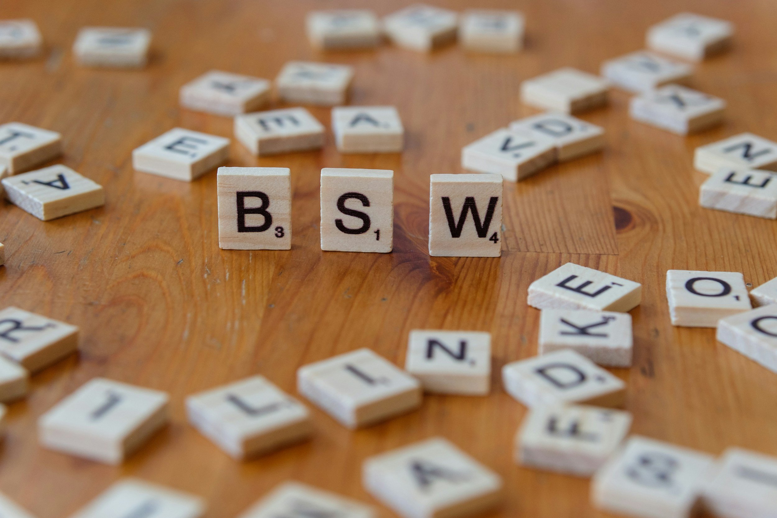 Scrabble tiles spelling BSW on a wooden surface surrounded by scattered letter tiles.