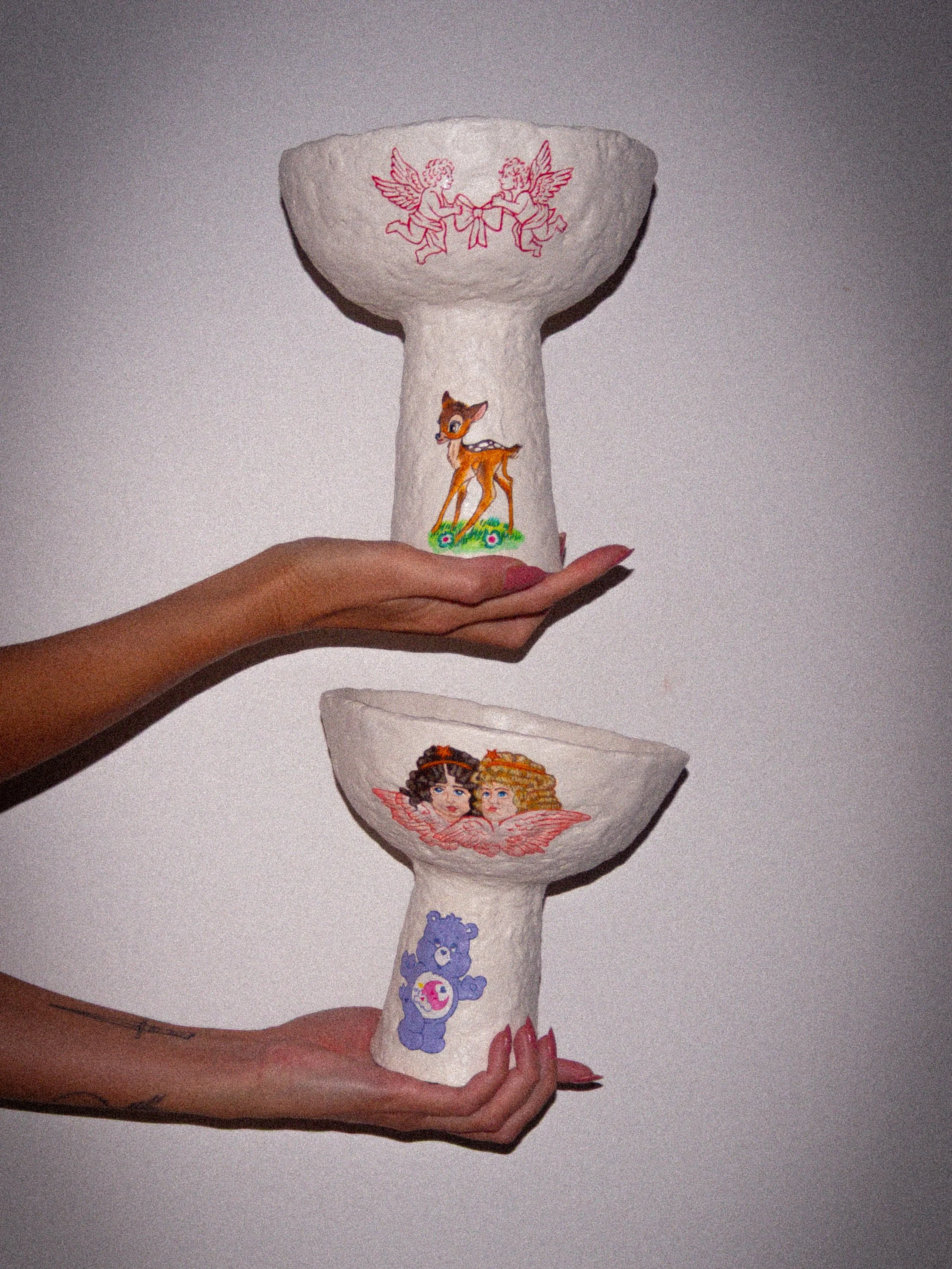 A photo of 2 sculptural bowls, made from paper pulp clay, hand painted with vintage and 1990s cartoons such as bambi, care bears, angels and cupids. the bowls are being held up by two female hands against a plain grey backdrop.