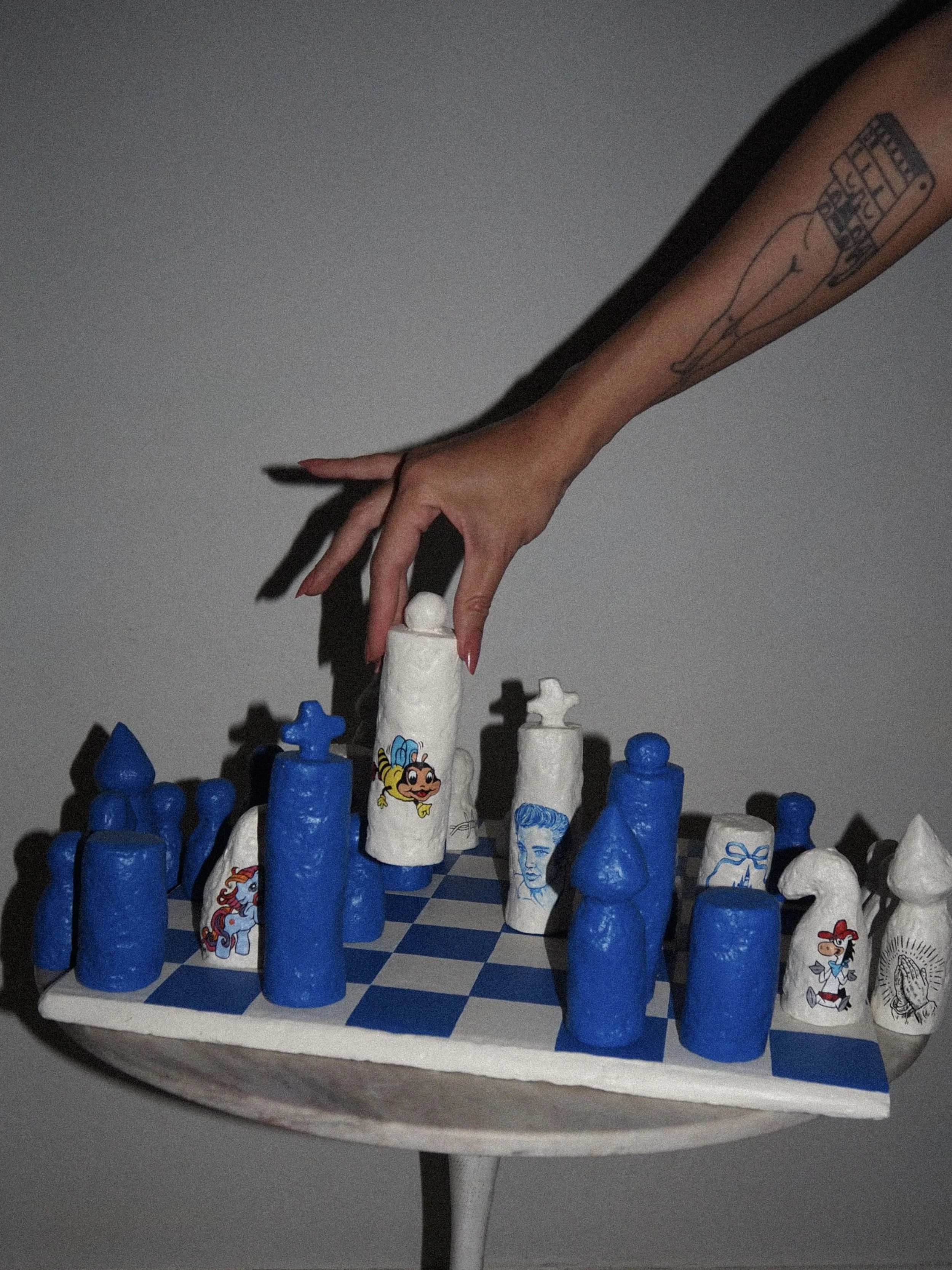 Close-up of a person's hand with a tattoo, reaching out to move a large chess piece with cartoon characters. The chessboard has alternating blue and white squares, and the pieces are decorated with drawings of characters like a mermaid, a woman, a penguin, and a unicorn. The scene is set against a plain background.
