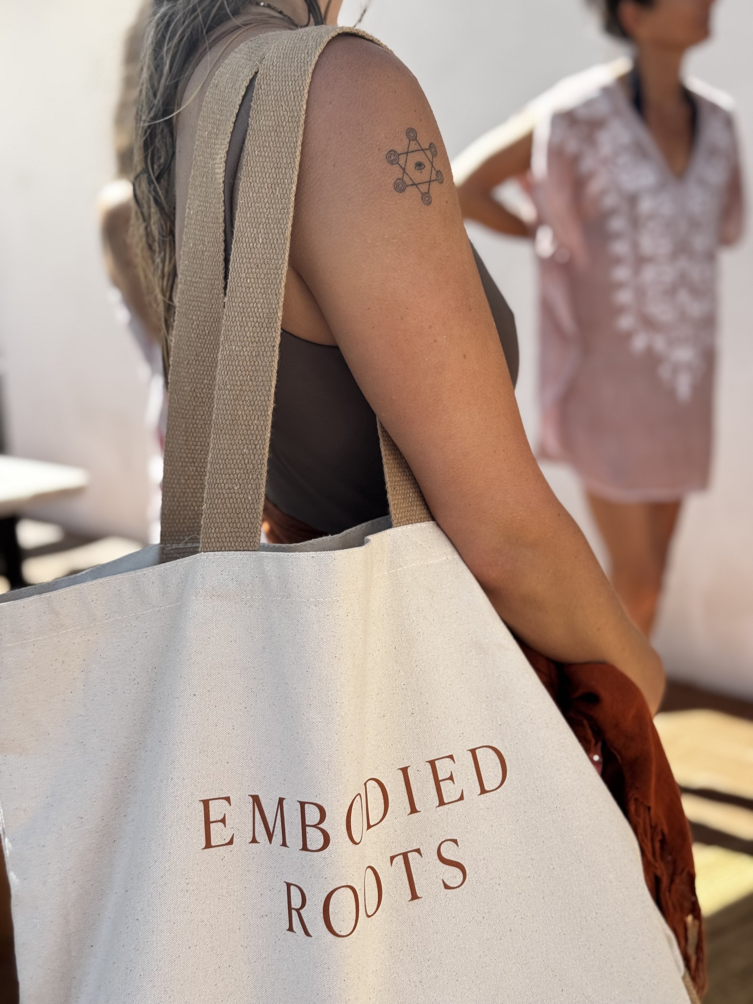 Close-up of a woman with a tattoo on her upper arm, carrying a beige tote bag with the words 'Embroidered Roots' from the yoga retreat printed on it, and another woman in the background wearing a pink embroidered outfit.