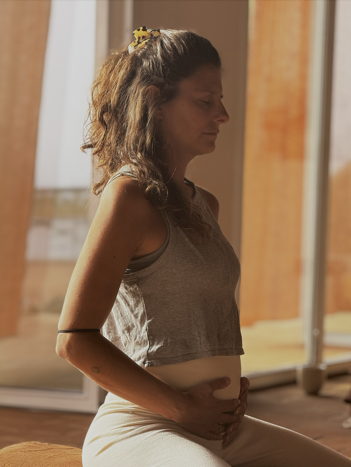 Sadie sitting, teaching pregnancy and pre-natal yoga with eyes closed, in a room with large windows and warm lighting.
