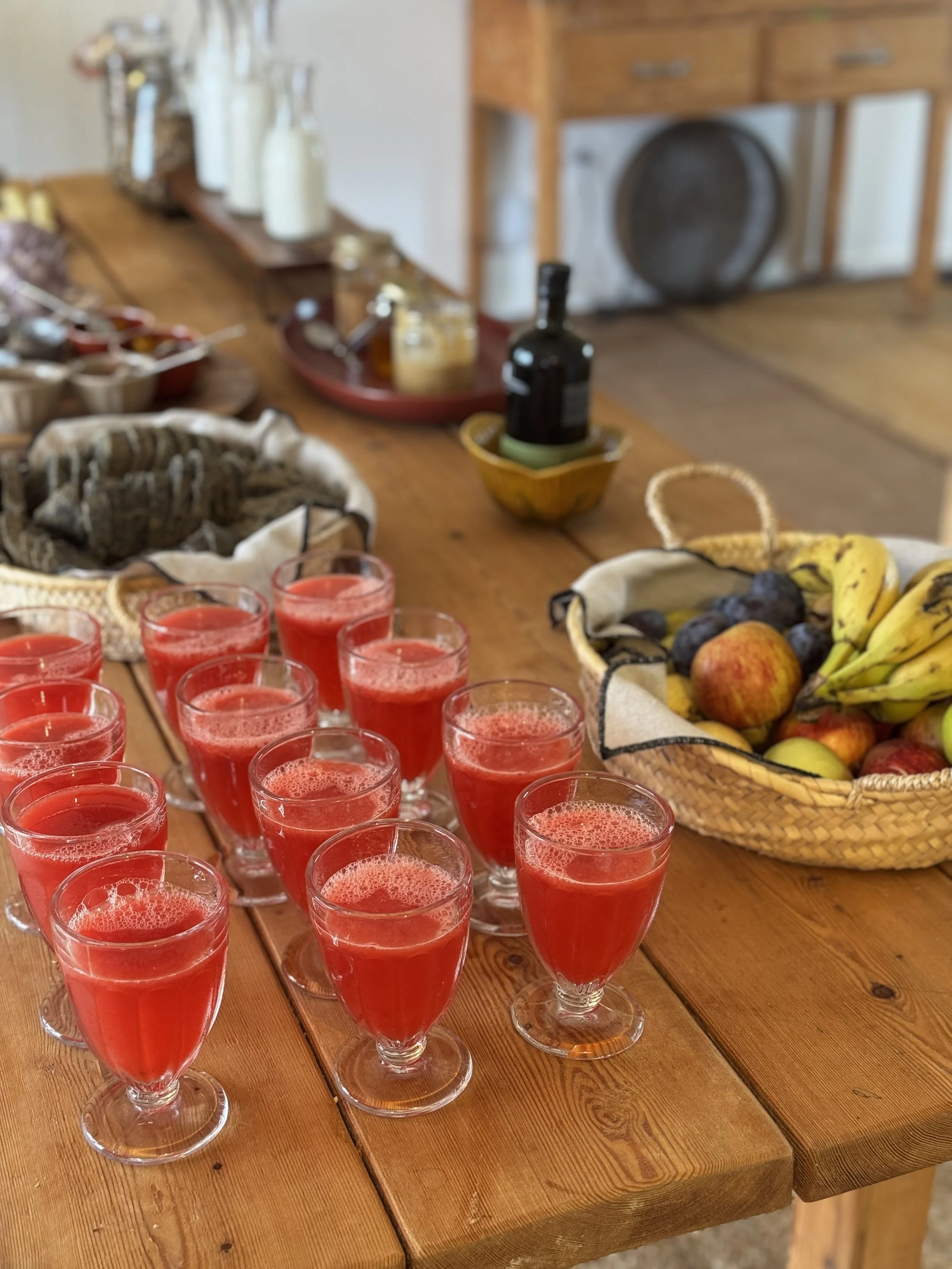A wooden table set with multiple glasses of red juice or beverage and a basket of assorted fresh fruits intended for yoga retreat guests in Portugal and Devon.