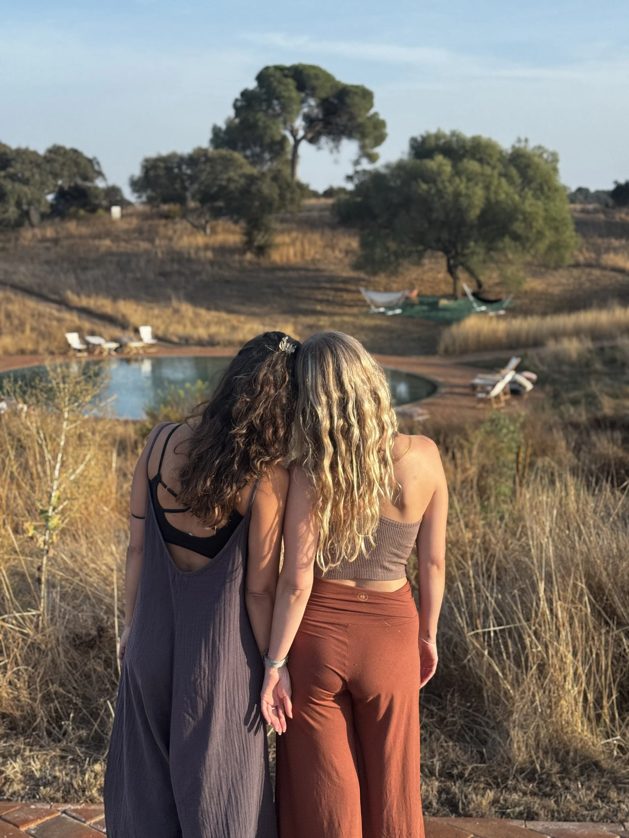 Two women stand close together outdoors overlooking a pool and landscape with trees. One woman has dark, curly hair, and the other has long, wavy blonde hair. They are celebrating being yoga retreat hosts in Portugal.