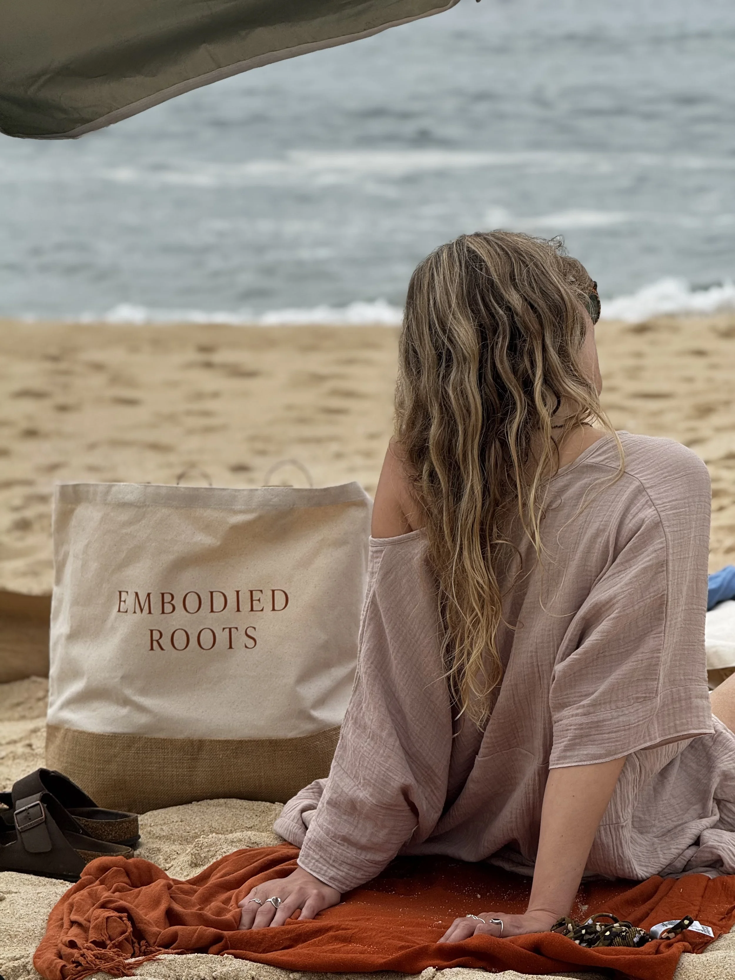 A person with long curly hair sitting on a beach at a yoga retreat in Portugal, with a bag labeled 'Embodied Roots' beside them, back facing the camera, near the shoreline with ocean waves in the background.